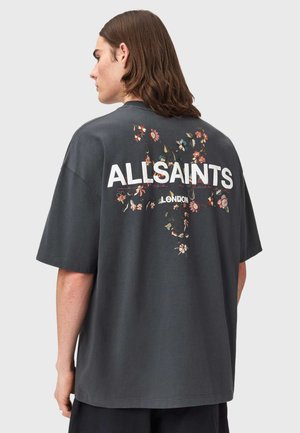 Hosszú barna hajú személy balra néz, sötétszürke, túlméretezett pólót visel, amelyen virágmintás kereszt és az "ALLSAINTS LONDON" felirat látható a hátoldalon.