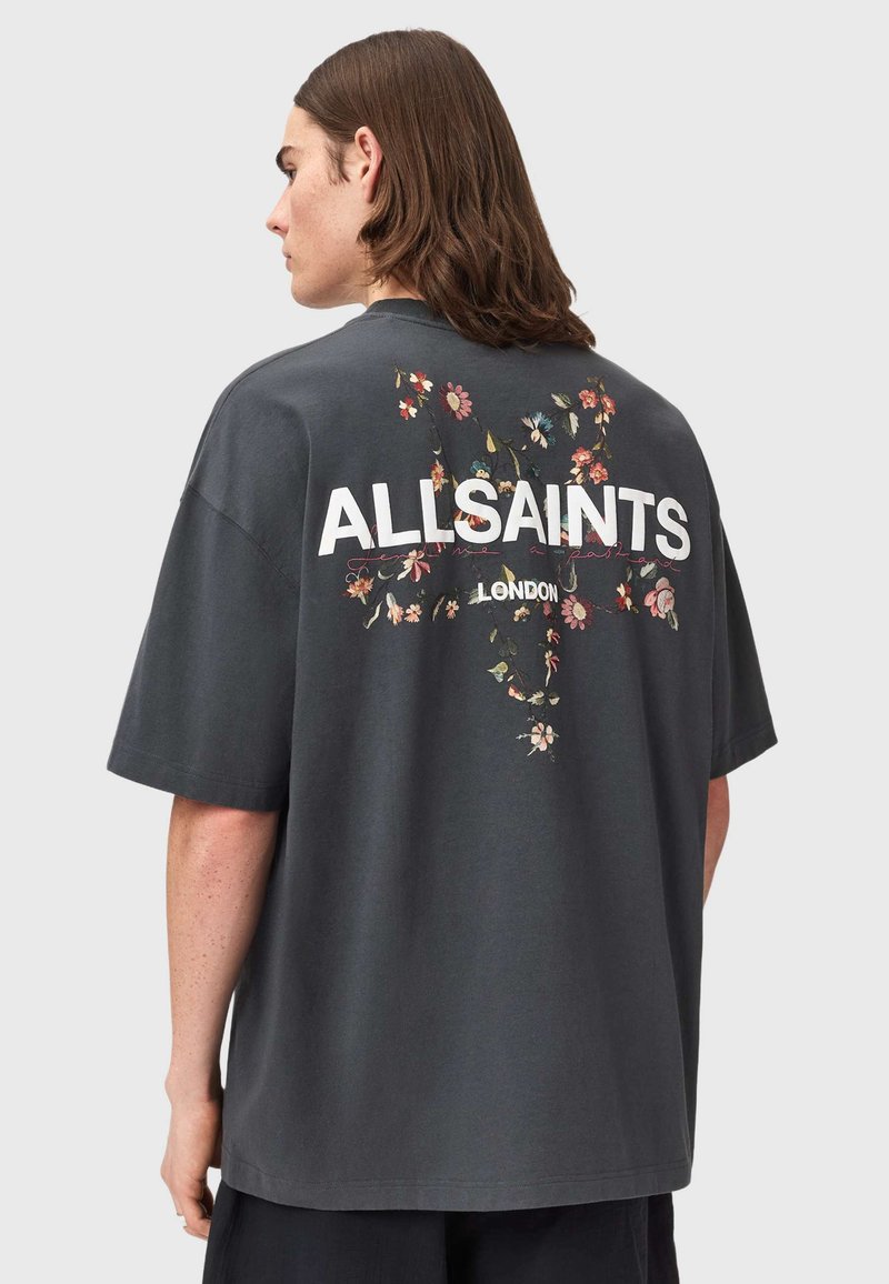 Άτομο με μακριά καστανά μαλλιά γυρισμένο προς τα αριστερά, φορώντας μια υπερμεγέθη σκούρα γκρι μπλούζα με σχεδιασμό λουλουδιών σε σχήμα σταυρού και το κείμενο "ALLSAINTS LONDON" στην πλάτη.