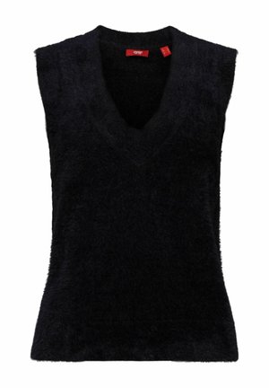 Femme portant une chemise blanche à boutons, un pull sans manches en tissu duveteux noir et une jupe en cuir noire, debout avec les bras partiellement levés.