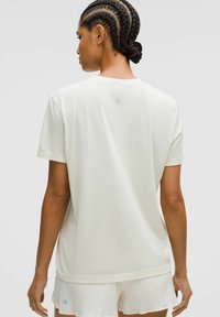 Vrouw met gevlochten haar die een witte T-shirt met korte mouwen en witte textuurshorts draagt, gezien van achteren tegen een effen achtergrond.