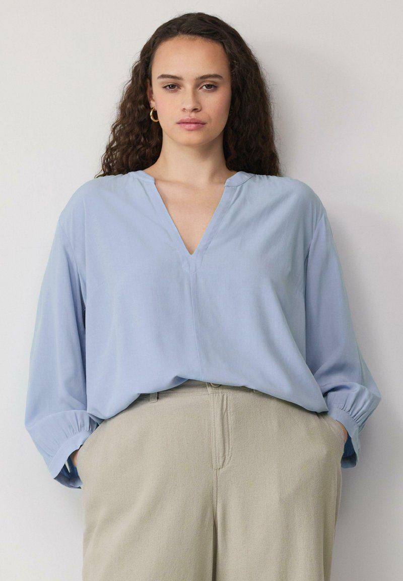 Lichtblauwe blouse met lange mouwen en een V-hals, voorzien van geplooide manchetten, gemaakt van gladde stof, gecombineerd met lichtbeige hoge taille broek.