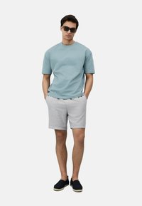 Light blue short-sleeve t-shirt paired with light gray shorts. Model wears dark sunglasses and black slip-on shoes with woven accents.