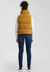 Frau trägt eine senfgelbe gepolsterte Weste, ein weißes Langarmshirt, dunkelblaue Skinny-Jeans und weiße Sneaker, mit dem Rücken zur Kamera.
