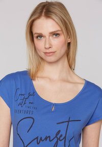 Blauwe katoen t-shirt met een ronde hals, korte mouwen en een sleutelgatdetail. Heeft zwarte tekstprints "Kom zeilen met mij" en "Zonsondergang."