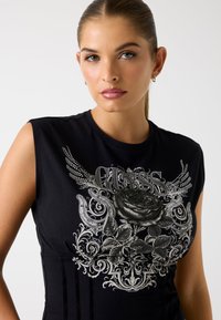 Top sin mangas de algodón negro con un diseño gráfico plateado que incluye una rosa, patrones decorativos y acentos de alas. Textura suave, diseño ajustado.