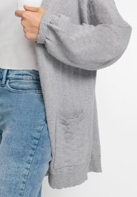 Světle šedý pletený cardigan s volným střihem, žebrovanými manžetami a malou kapsou na přední straně. Nosený přes bílé tričko a modré džíny.