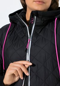 Giacca trapuntata nera con chiusura frontale con zip, zip bianca a contrasto e coulisse rosa acceso sul cappuccio. Dettagli in tessuto testurizzato sono visibili.