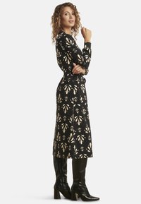 Schwarzes, langärmliges Kleid mit cremefarbenen Blumenmustern, tailliertem Schnitt und A-Linien-Silhouette, kombiniert mit knielangen schwarzen Stiefeln mit Absatz.