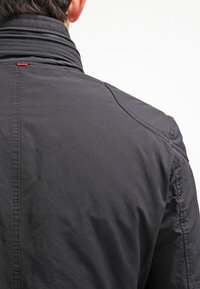 Svart jacka med hög krage, i en texturerad tyg, med synliga sömmar och en liten röd accent nära kragsömmen.