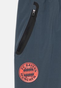 Bayern Münchens treningbukser i mørkeblåt stoff. Har en svart glidelåslomme og en rund rød logo med et særpreget triangelmønster.