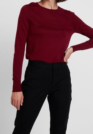 Femme portant un pull bordeaux à manches longues rentré dans un pantalon noir taille haute, debout avec une main sur la hanche et l'autre détendue.