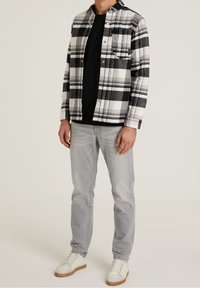 Geruit flanellen overhemd in zwart-wit, met een knoopsluiting aan de voorkant en een zak. Draag het over een zwart T-shirt, gecombineerd met lichtgrijze jeans en witte sneakers.