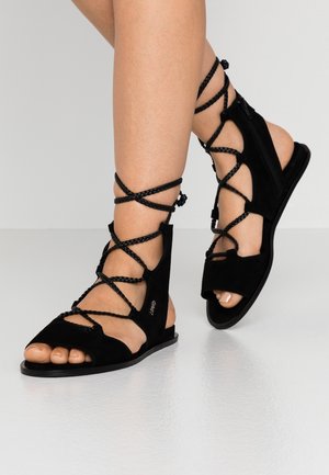 Jambes portant des sandales gladiateur plates noires avec des lanières tressées enroulées autour des chevilles et une fermeture éclair latérale.