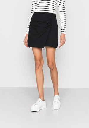 Frau trägt einen schwarzen, gerafften Minirock, weiße Plateausneaker und ein langärmliges weißes Hemd mit schwarzen horizontalen Streifen.