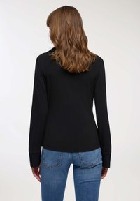 Donna con i capelli castani di lunghezza media, che indossa una maglietta nera a maniche lunghe e jeans blu, di spalle su uno sfondo chiaro e semplice.