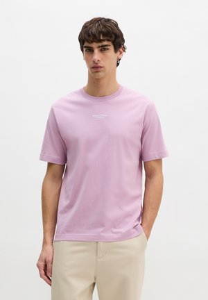 Jonge man die een lichtpaarse Marc O’Polo T-shirt en beige broek draagt, staand tegen een effen witte achtergrond.