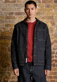 Dark denim jacket with a classic collar, featuring two front pockets and contrasting stitching, layered over a red sweatshirt.
