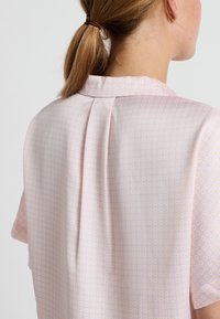 Blusa a maniche corte in tessuto rosa chiaro con un sottile motivo floreale su tutta la superficie e un dettagli di piega sul retro. Texture morbida e liscia.