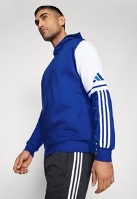 Blaues Kapuzensweatshirt mit weißen Ärmeln und drei diagonalen Streifen. Das Material scheint weich zu sein, mit einem lockeren Schnitt und einer Kängurutasche.