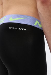 Κοντινή λήψη ενός ατόμου που φοράει μαύρο κολάν Nike Dri-FIT ADV με μωβ ζώνη μέσης που φέρει πράσινο-λαχανί λογότυπο Nike.