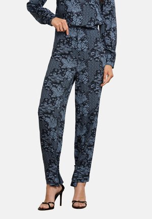Donna con pantaloni blu scuro a motivi floreali e top a maniche lunghe coordinato, che indica la vita, in piedi con sandali neri con tacco alto.