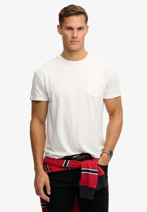 Uomo che indossa una t-shirt bianca a maniche corte con una tasca sul petto, pantaloni neri e un maglione a righe rosse legato in vita.