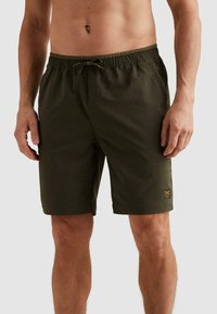 Buste masculin portant un short taille élastique vert foncé avec cordon de serrage et petite étiquette logo sur la jambe gauche, debout devant un fond uni.