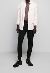 Camisa de botón color beige claro sobre una camiseta marrón, combinada con pantalones negros y botas de tobillo negras y brillantes. La camisa tiene un bolsillo en el pecho.