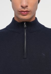 Navyblauer gestrickter Reißverschluss-Pullover mit hohem Kragen, geripptem Textur und dezentem Logodetail in der Mitte am Saum.