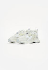 Baskets Lacoste en beige clair avec des panneaux texturés, des accents gris et une semelle épaisse. Présente un branding visible et un système de laçage.