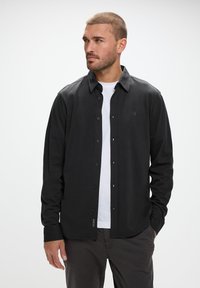 Svart skjorta med knappar i mjukt tyg, med klassisk krage, långa ärmar och en subtil logotyp på vänster bröst.