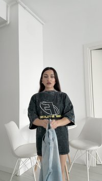 Przesadny czarny T-shirt z białą i żółtą grafiką, trzymany w jasnoniebieskich dżinsach; nowoczesne wnętrze z białymi krzesłami w tle.