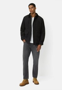 Schwarze Wollmischung Jacke mit vier Vordertaschen, graue Jeans und beigefarbene Schnürstiefel. Darunter ein weißes T-Shirt. Modell steht vor einem weißen Hintergrund.
