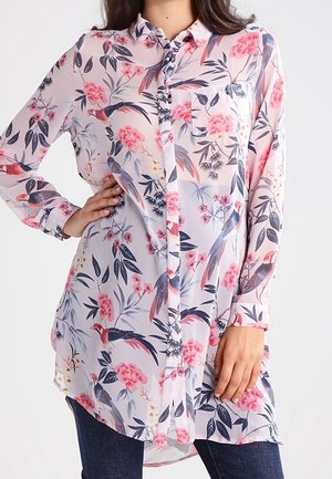 Langærmet knapbluse med pink og røde blomster- og fuglemønstre, båret over mørkeblå jeans, modelens hånd på hoften og siden.