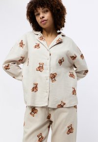 Linnedrakt settet har beige stoff med brune teddy-bjørn illustrasjoner. Knappeløs skjorte har en avslappet passform; buksene matcher med gjentatt mønster.