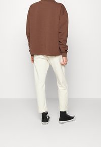 Felpa marrone oversize, pantaloni color crema chiaro e sneakers alte nere. Materiale in cotone con vestibilità comoda e polsini arrotolati sui pantaloni.