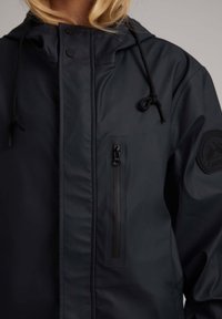 Veste marine résistant à l'eau avec une poche poitrine zippée, une capuche à cordon de serrage et un tissu texturé avec un patch logo sur la manche.
