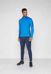 Niebieska sportowa bluza z długim rękawem i czarnymi akcentami, z zamkiem na szyi, w połączeniu z granatowymi dopasowanymi spodniami i czarnymi butami piłkarskimi z niebieskimi detalami.