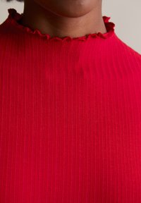 Rotes, geripptes Oberteil mit hohem Kragen und rüschendetail. Der Stoff hat eine glatte Textur und ein figurbetontes Design.