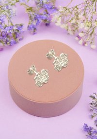 Silverblommiga örhängen med en texturerad yta, monterade på en rund, blush pink sammetshållare. Omgiven av små lila och vita blommor.