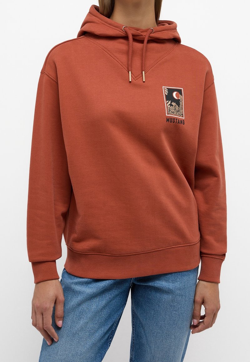 Sudadera de color óxido con parche gráfico floral y lunar en el frente, cordones con punta dorada, llevada con jeans de mezclilla azul por una persona de pie.