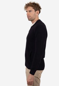 Suéter de punto azul marino oscuro con cremallera, con bolsillos laterales y un patrón texturizado, usado sobre una camisa blanca, combinado con pantalones beige.