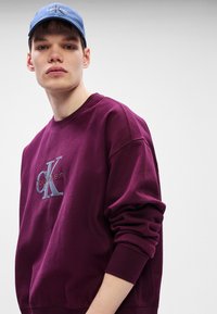 Bordový svetr s kulatým výstřihem, na kterém je vyšitý logo "Calvin Klein" v šedé a modré barvě. Měkká textura, volný střih.