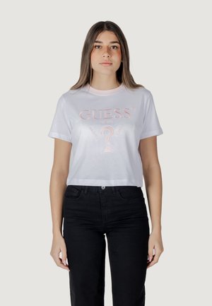 Camiseta blanca corta con el logo bordado en rosa "GUESS" y un signo de interrogación. Mangas cortas; combinada con unos vaqueros de denim negros.