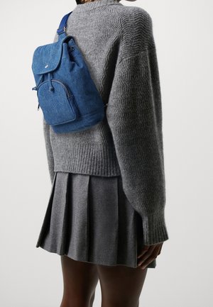 Persoon draagt een blauwe denim rugzak, een grijze gebreide trui met textuur en een geplooide grijze rok, gezien vanaf de achterkant tegen een eenvoudige achtergrond.