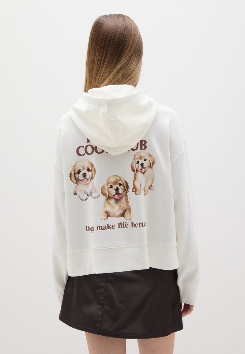 Crème hoodie met een trekkoordkap, met drie cartoonhonden en de tekst "Honden maken het leven beter" op de achterkant. Gecombineerd met een zwarte rok.