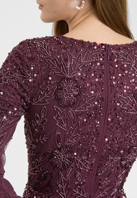 Abito borgogna con dettagli intricati in perline e paillettes, ricami floreali e un tessuto testurizzato. Presenta una zip nascosta sul retro.