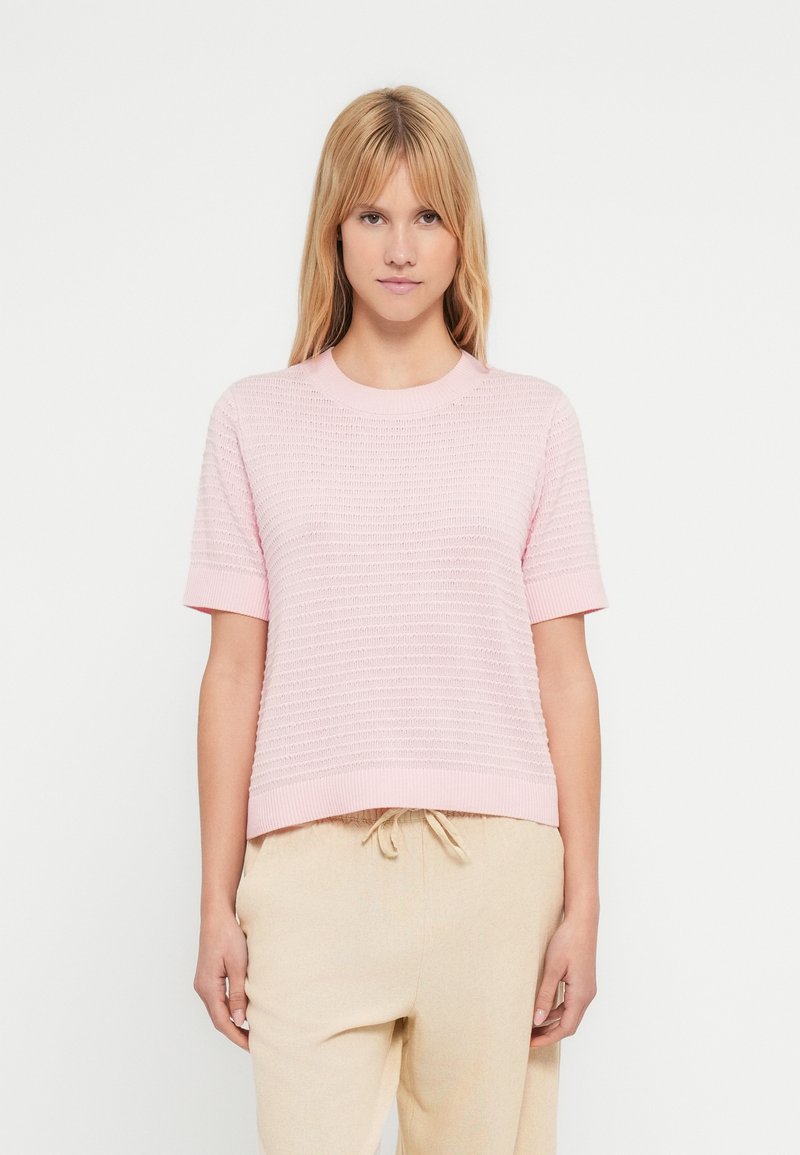 Pull rose clair en tricot avec un motif texturé, col rond et manches courtes, associé à un pantalon beige à taille élastique.