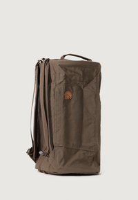 Mochila de tela marrom com formato retangular, puxador na parte de cima, alças ajustáveis e um emblema. Possui um compartimento principal com fecho de correr e um bolso lateral.