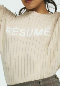 Beige geribbelde trui met witte tekst "RÉSUMÉ" op de borst. Zacht gebreid materiaal, ronde halslijn en zichtbare naden voegen textuur toe.